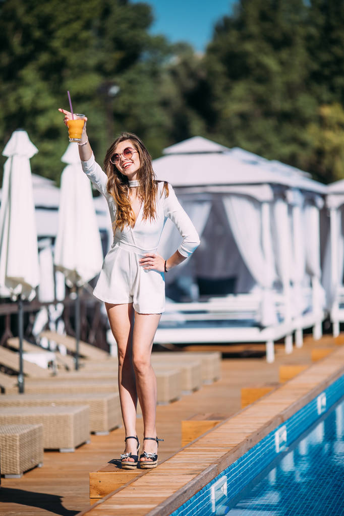 uśmiechnięta emocjonalna dziewczyna nosi białą modną letnią sukienkę pozującą obok egzotycznej letniej plaży. Niesamowita kobieta pije koktajle w pobliżu basenu.party. - Zdjęcie, obraz
