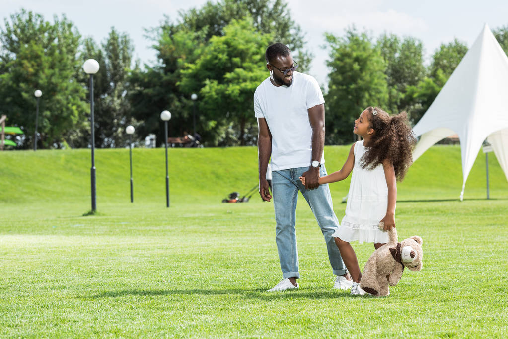 Afrikaanse Amerikaanse vader en dochter met teddy bear hand in hand en wandelen in het park - Foto, afbeelding