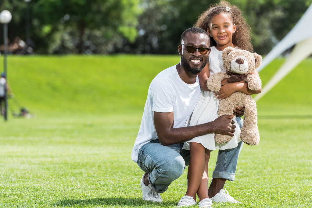 africano americano padre y hija con teddy oso mirando cámara en parque
 - Foto, Imagen