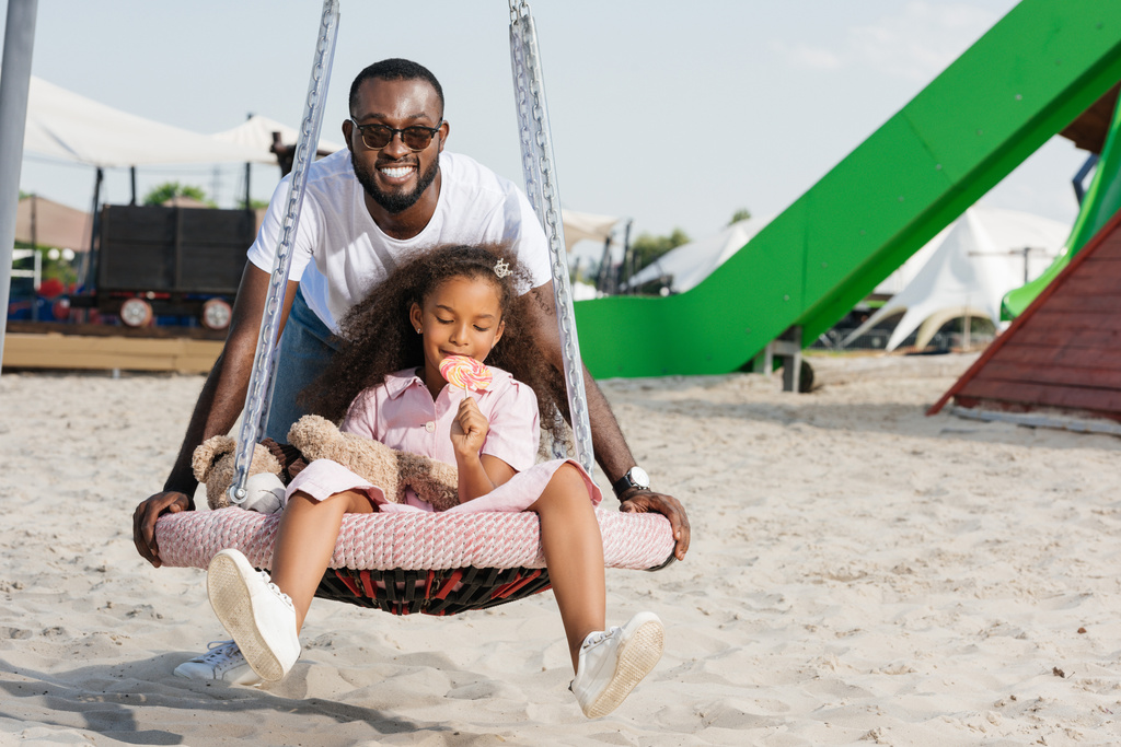 sorridente padre afroamericano spingendo figlia su ragnatela nido altalena con lecca-lecca al parco giochi
 - Foto, immagini