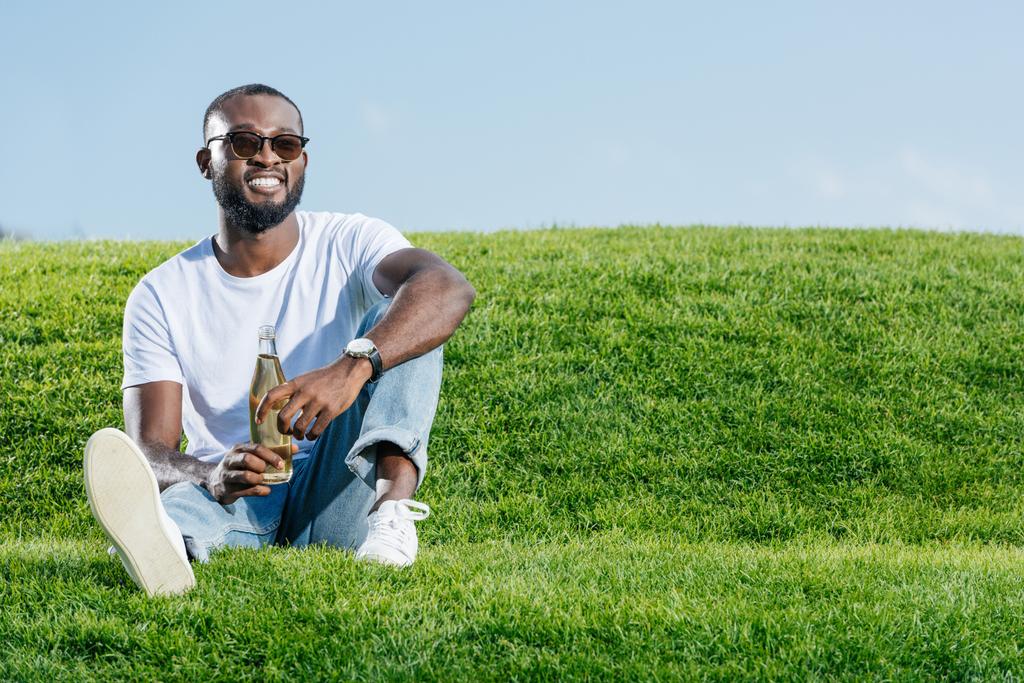 hombre afroamericano guapo feliz sentado con soda en botella de cristal en la colina verde
 - Foto, Imagen