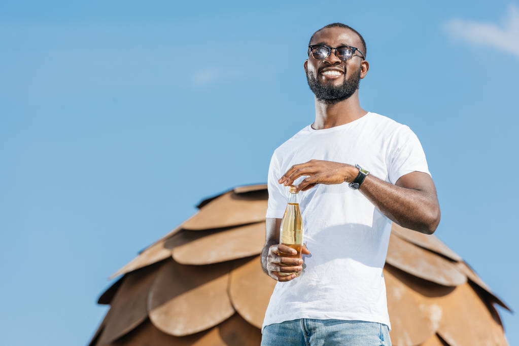 uśmiechający się przystojny mężczyzna african american, otwierając butelkę soda przeciw błękitne niebo - Zdjęcie, obraz
