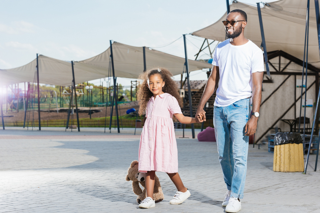 χαμογελώντας αφρικανικό αμερικανικό πατέρα και κόρη, κρατώντας τα χέρια και τα πόδια στο λούνα παρκ - Φωτογραφία, εικόνα