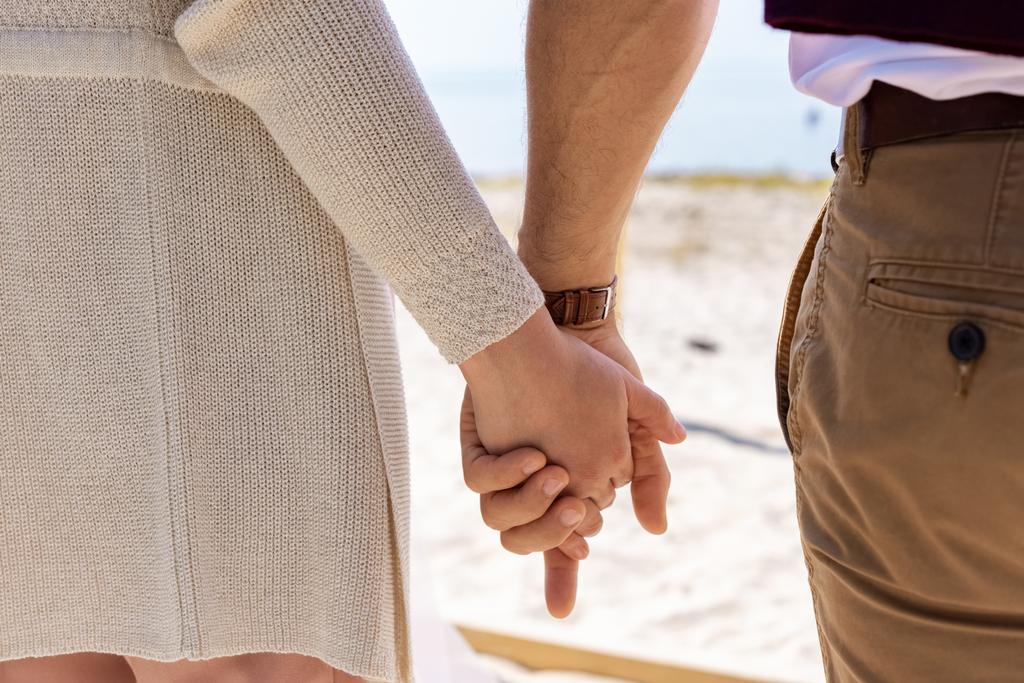 砂浜のビーチを歩きながら手を繋いでいるカップルの部分的なビュー - 写真・画像