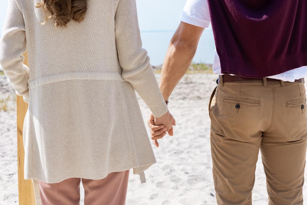 částečný pohled páru, drželi se za ruce při chůzi na písečné pláži - Fotografie, Obrázek