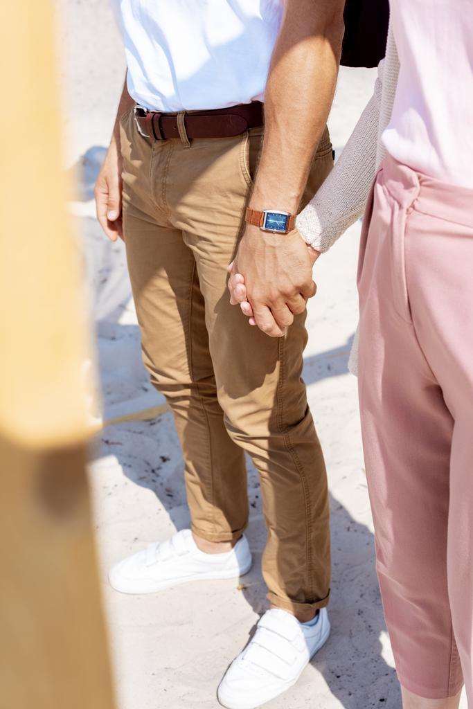 частичный вид пары, держащейся за руки во время прогулки по песчаному пляжу
 - Фото, изображение