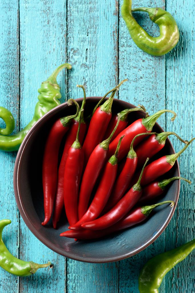 pohled shora červené chilli papričky v misce a rozptýlené zelené papriky na modré dřevěný stůl - Fotografie, Obrázek