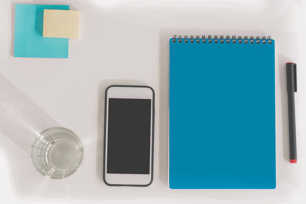 Верхний вид смартфона с чистым экраном, ноутбук с ручкой, стакан воды и липкие ноты
 - Фото, изображение