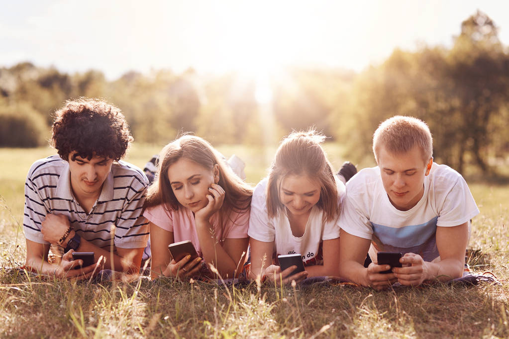 Four friends focused in their smart phones, enjoy leisure time on nature, lie on green grass, share photos after picnic in their social networks, pose against sunshine. Teenagers, technology - Photo, Image
