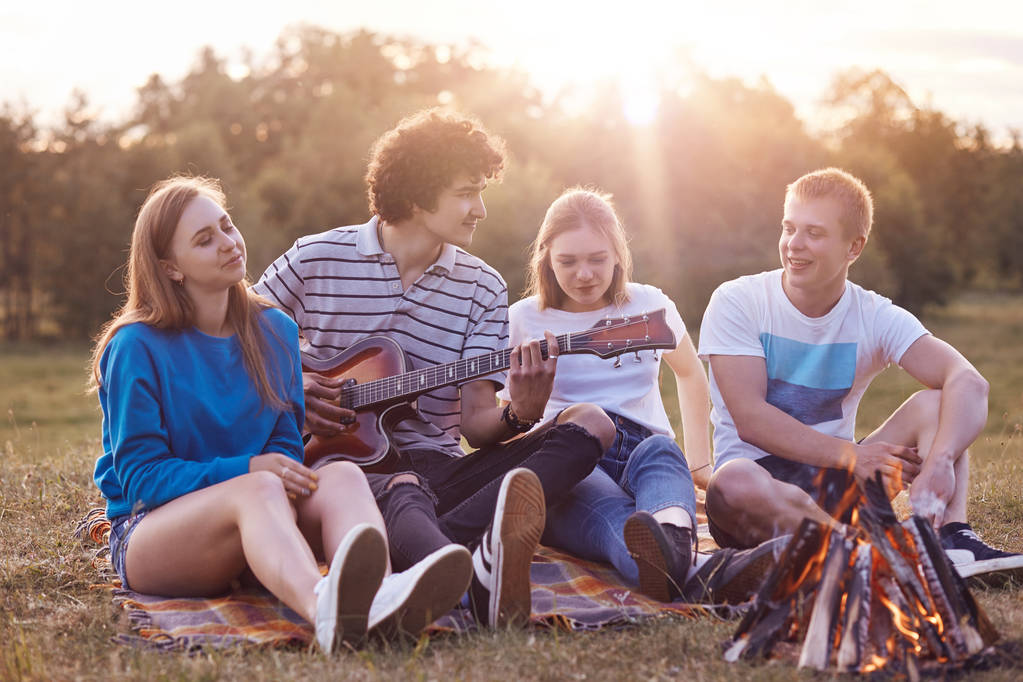 People and vacation concept. Cheerful best teenage friends enjoy romantic atmosphere on nature, have picnic together, play guitar, sit on plaid near campfire, have friendly talk with each other - Photo, Image