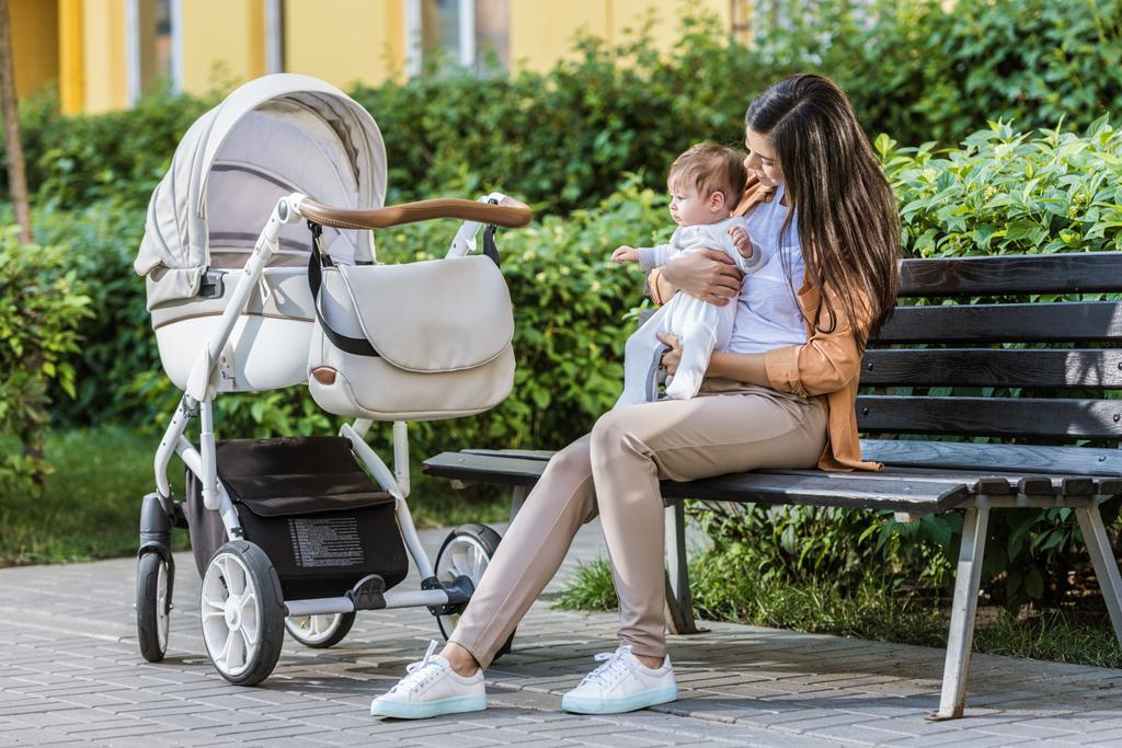 mother sitting with baby on bench near stroller in park - Photo, Image