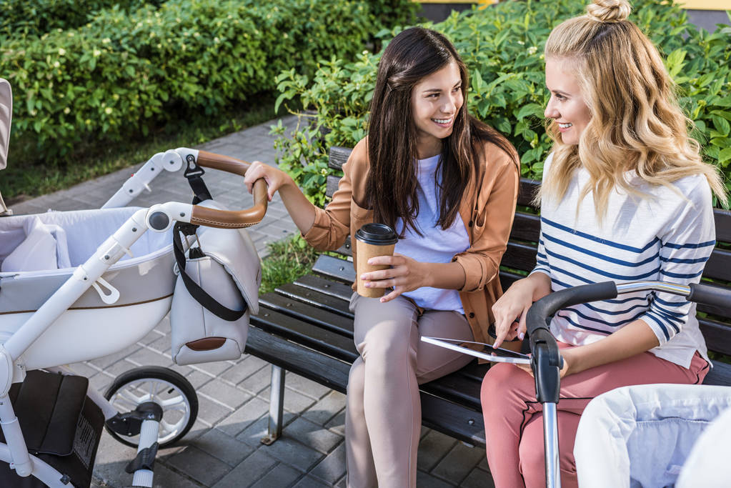 madres sonrientes sentadas en el banco cerca de cochecitos de bebé con café en taza de papel y tableta digital
 - Foto, Imagen