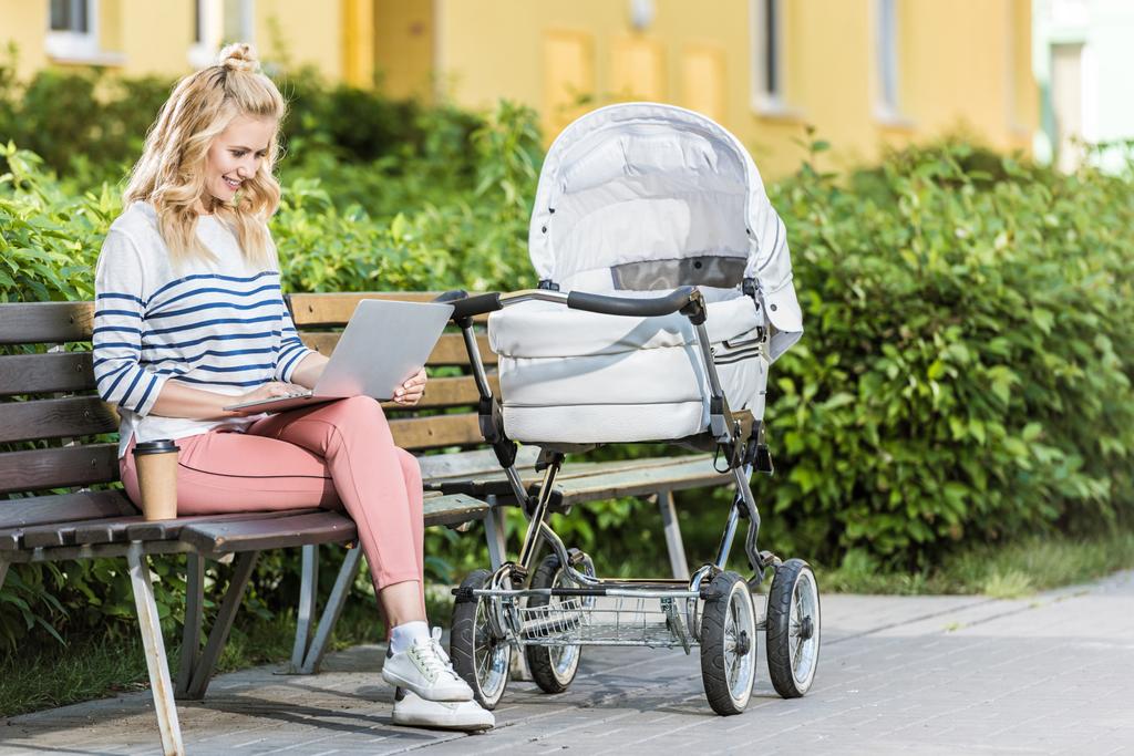 freelancer sorrindo trabalhando com laptop no banco perto de carrinho de bebê no parque
 - Foto, Imagem