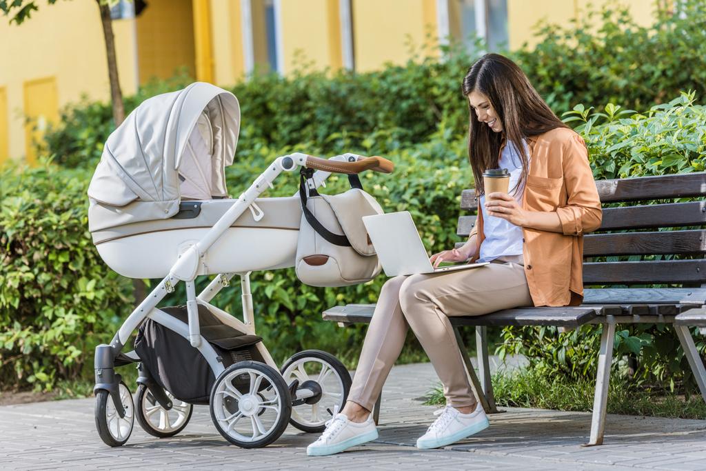 pigiste travaillant avec un ordinateur portable sur un banc près de poussette bébé dans le parc et tenant du café dans une tasse en papier
 - Photo, image