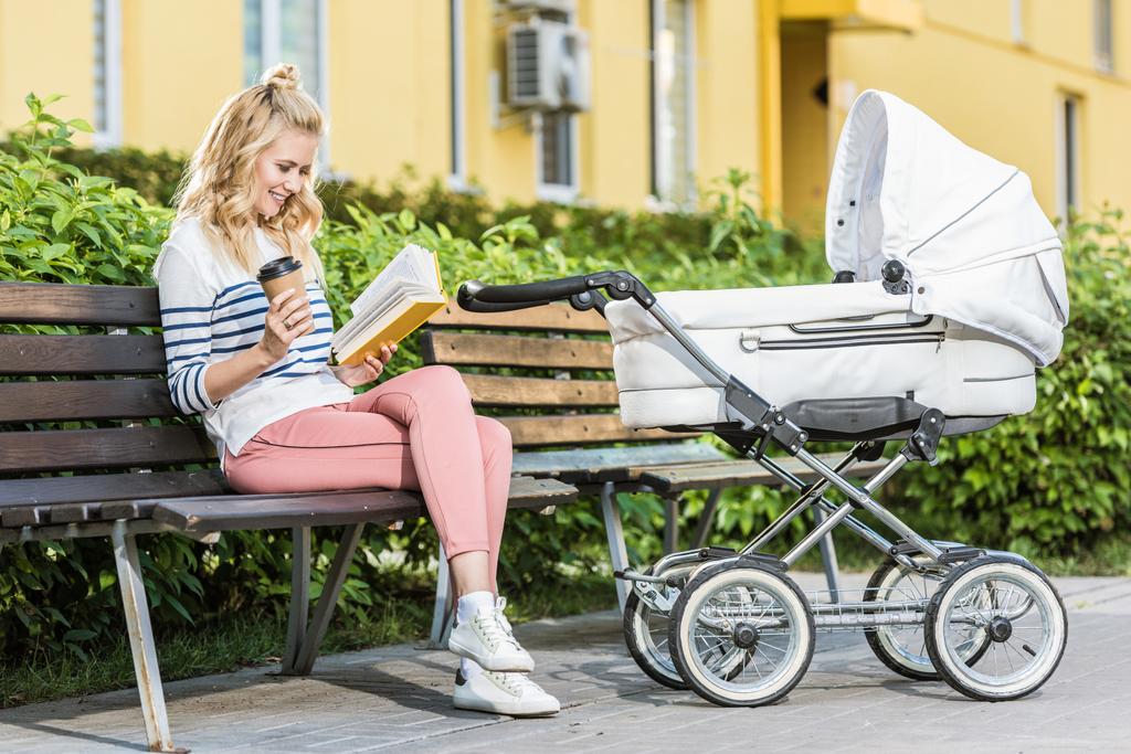 glimlachend moeder leesboek en houden van koffie te gaan op de Bank in de buurt van baby wandelwagen in park - Foto, afbeelding