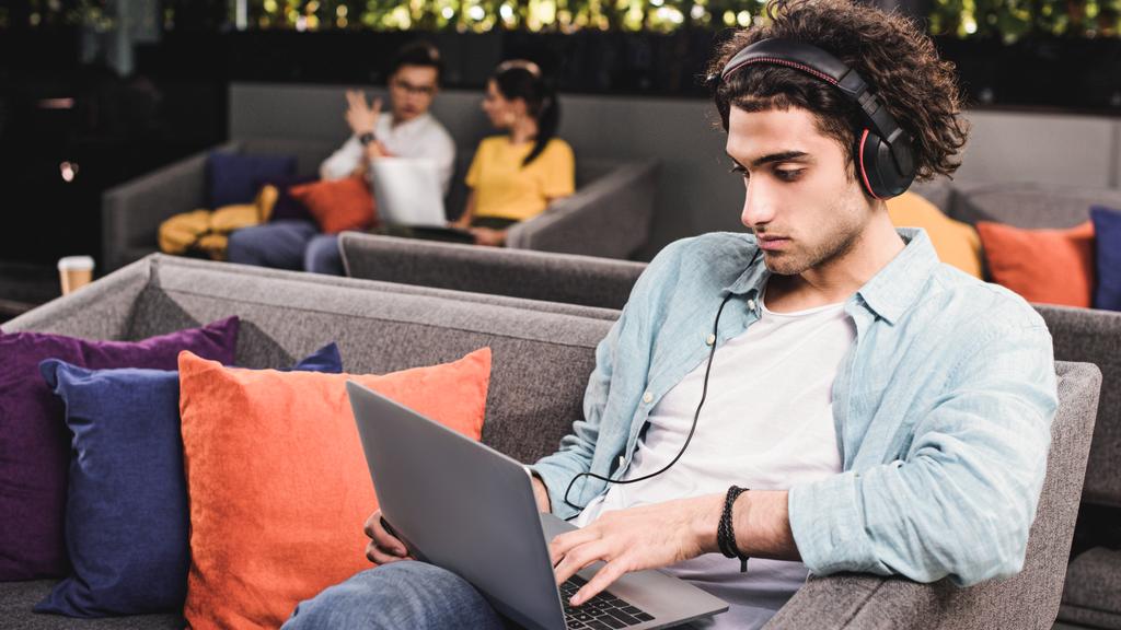 молодий бізнесмен в навушниках, сидячи на дивані з ноутбуком в сучасному коворкінгу
  - Фото, зображення