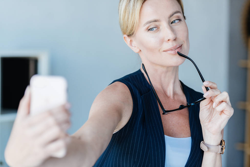 schöne erwachsene Geschäftsfrau mit Brille macht Selfie auf Smartphone im Büro  - Foto, Bild