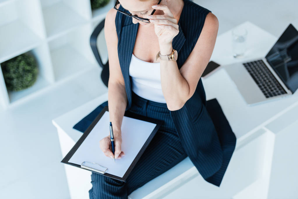 vista de ángulo alto de la mujer de negocios escribiendo en portapapeles en la mesa con el ordenador portátil en la oficina
  - Foto, Imagen