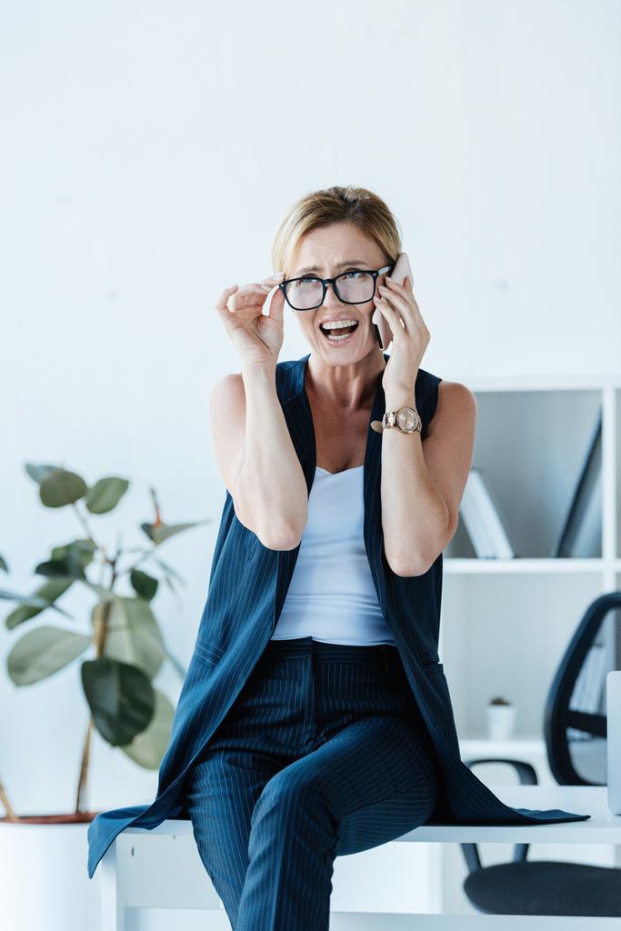emotional businesswoman touching eyeglasses and talking on smartphone in office  - Photo, Image