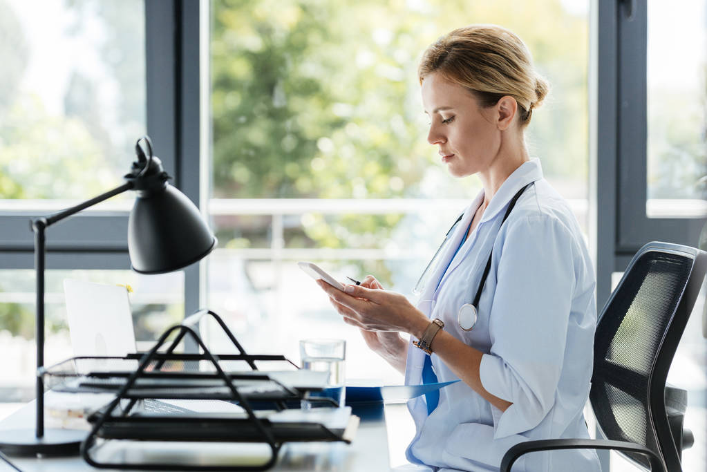 sivunäkymä naislääkärin valkoisessa takissa käyttäen älypuhelin pöydässä toimistossa
  - Valokuva, kuva
