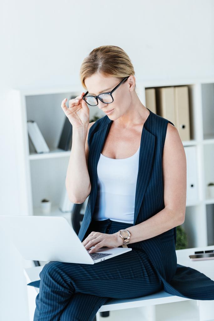 Konzentrierte Geschäftsfrau mit Brille sitzt am Tisch und arbeitet im Büro am Laptop  - Foto, Bild