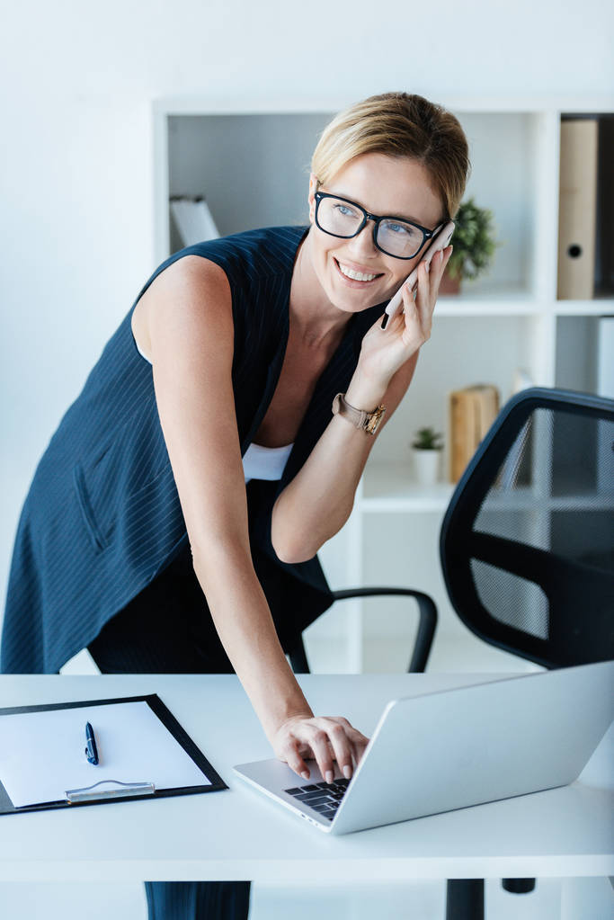 erwachsene Geschäftsfrau mit Brille spricht auf Smartphone und benutzt Laptop im Büro  - Foto, Bild