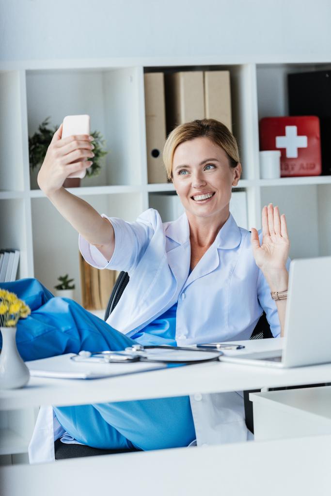 glückliche Ärztin mit Videoanruf auf dem Smartphone und Handwinken im Büro  - Foto, Bild