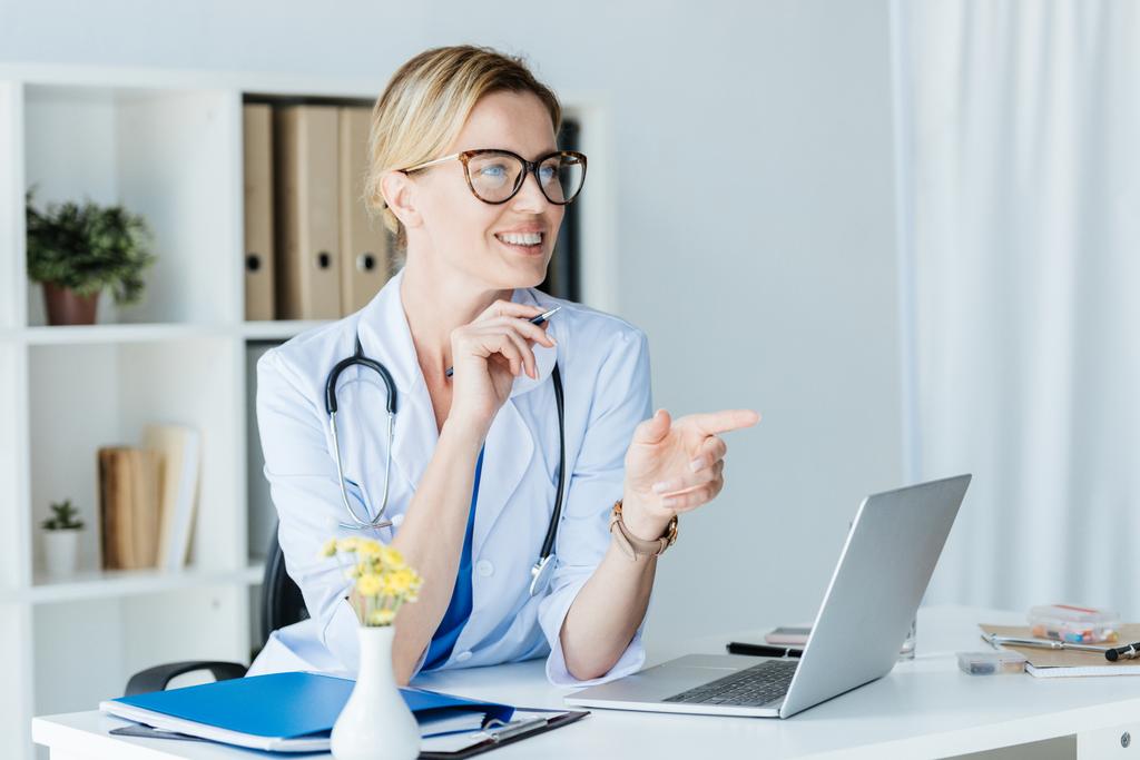 улыбающаяся женщина-врач в очках, указывающая пальцем и говорящая за столом с ноутбуком в офисе
  - Фото, изображение