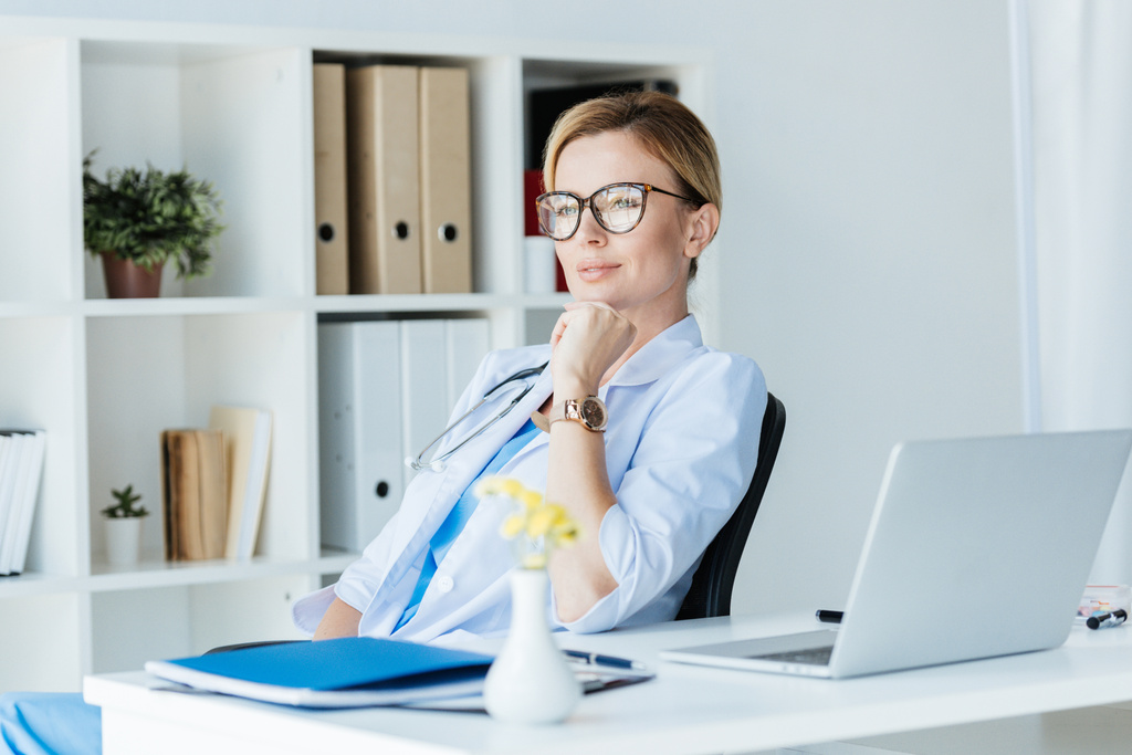 συλλογισμένος ενήλικη γυναίκα γιατρό σε γυαλιά, κάθεται στο τραπέζι με το laptop στο γραφείο  - Φωτογραφία, εικόνα