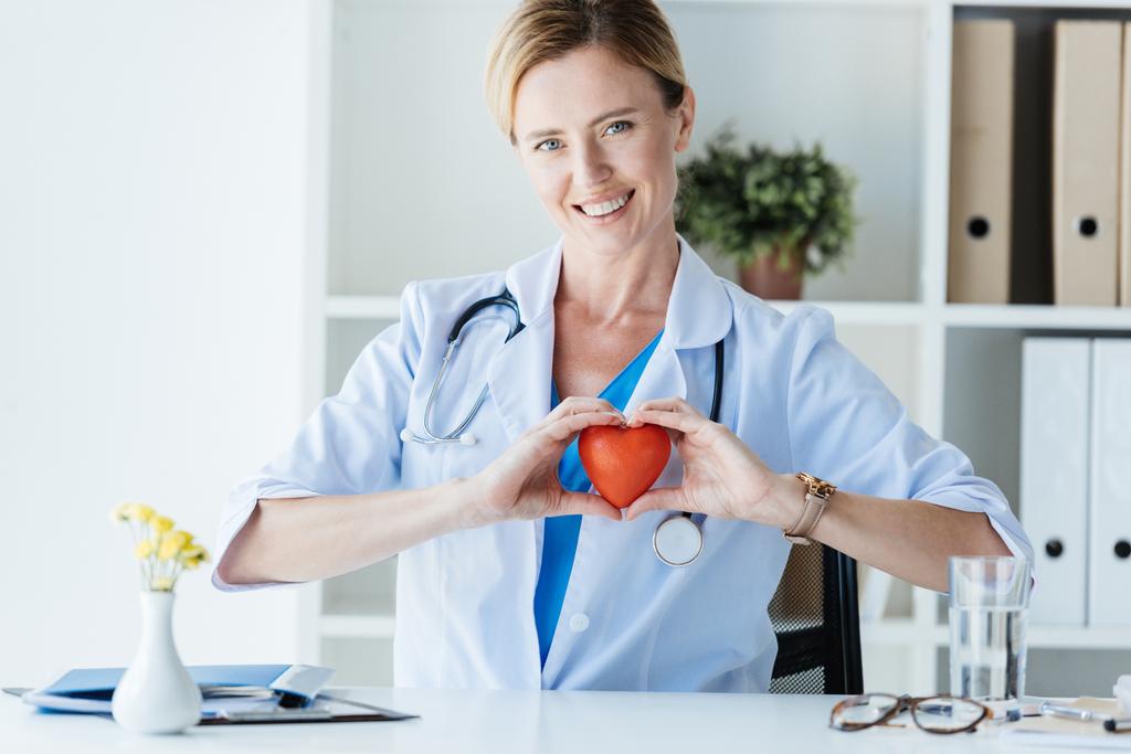 ευτυχισμένη γυναίκα γιατρό σε λευκό παλτό δείχνει το σύμβολο καρδιά στο τραπέζι στο γραφείο  - Φωτογραφία, εικόνα
