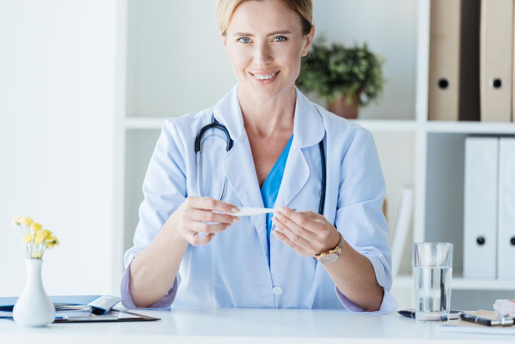attraktive erwachsene Ärztin im weißen Mantel mit Thermometer am Tisch im Büro  - Foto, Bild