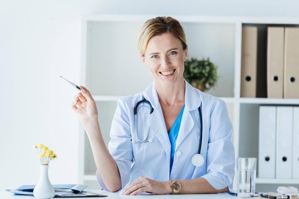 улыбающаяся женщина-врач в белом халате показывает термометр за столом в офисе
  - Фото, изображение