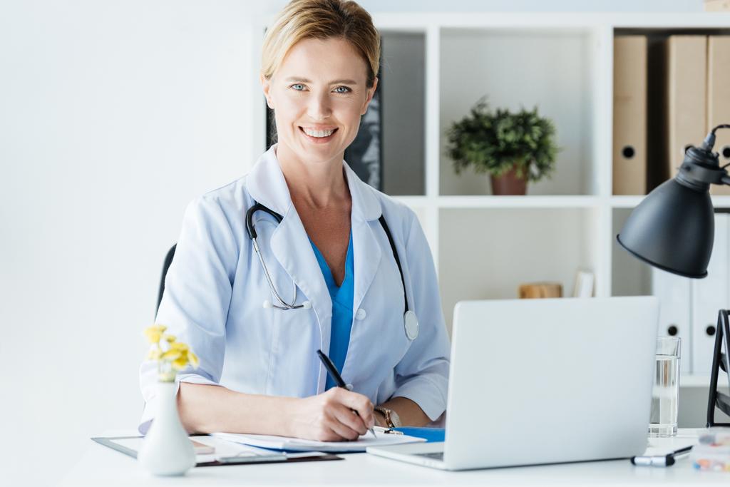 adult female doctor with stethoscope over neck writing in clipboard at table with laptop in office  - Photo, Image