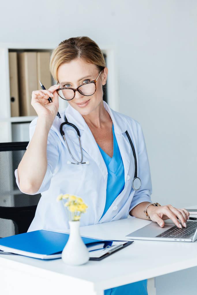 attraktive erwachsene Ärztin mit Brille, die in die Kamera schaut, während sie am Laptop am Tisch im Büro arbeitet - Foto, Bild