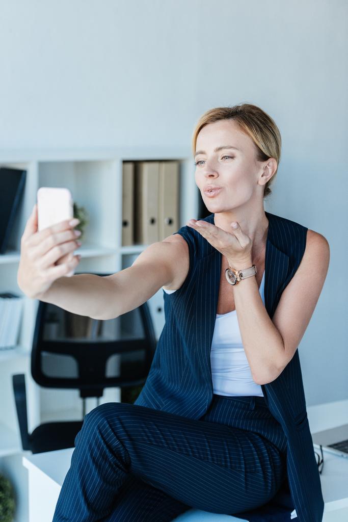 オフィスでスマート フォンでビデオ通話中に空気キスを吹いている魅力的なアダルト女性実業家  - 写真・画像
