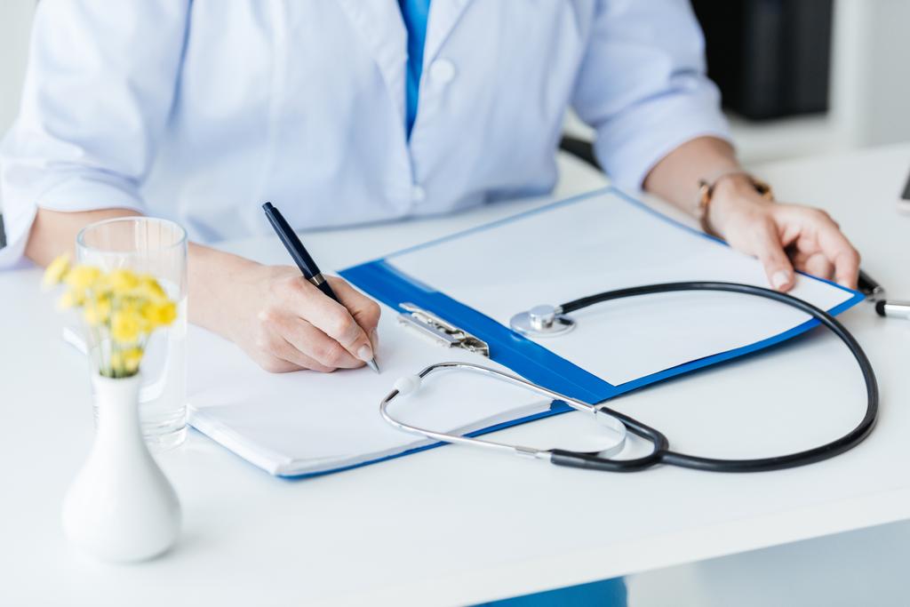 cropped image of female doctor writing in clipboard at table with stethoscope in office  - Photo, Image
