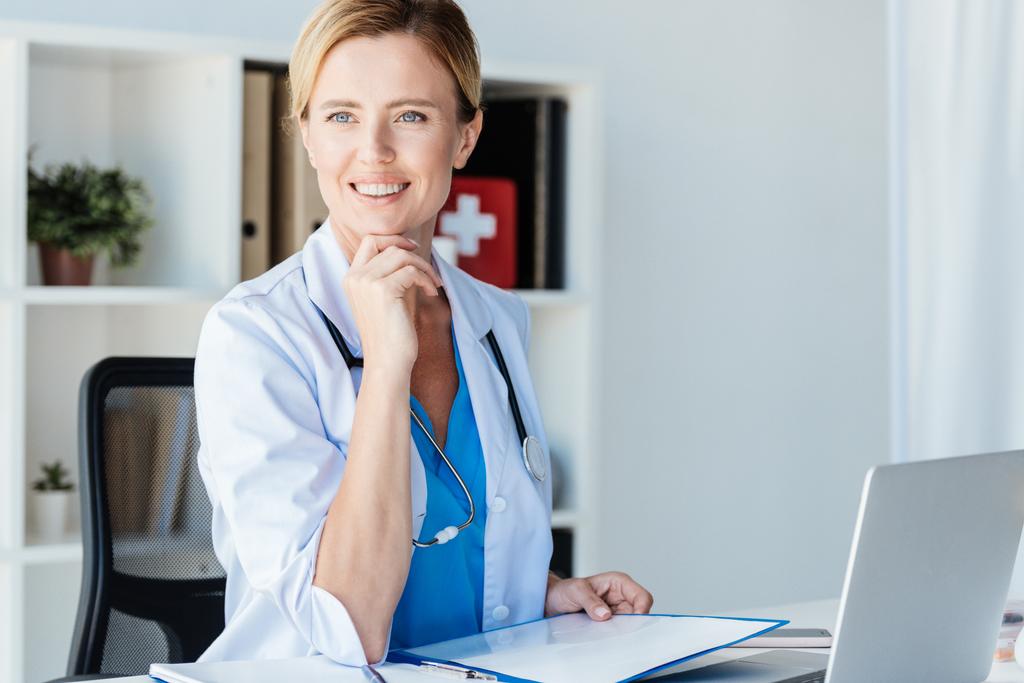 улыбающаяся женщина-врач со стетоскопом смотрит в сторону стола с буфером обмена и ноутбуком в офисе
  - Фото, изображение