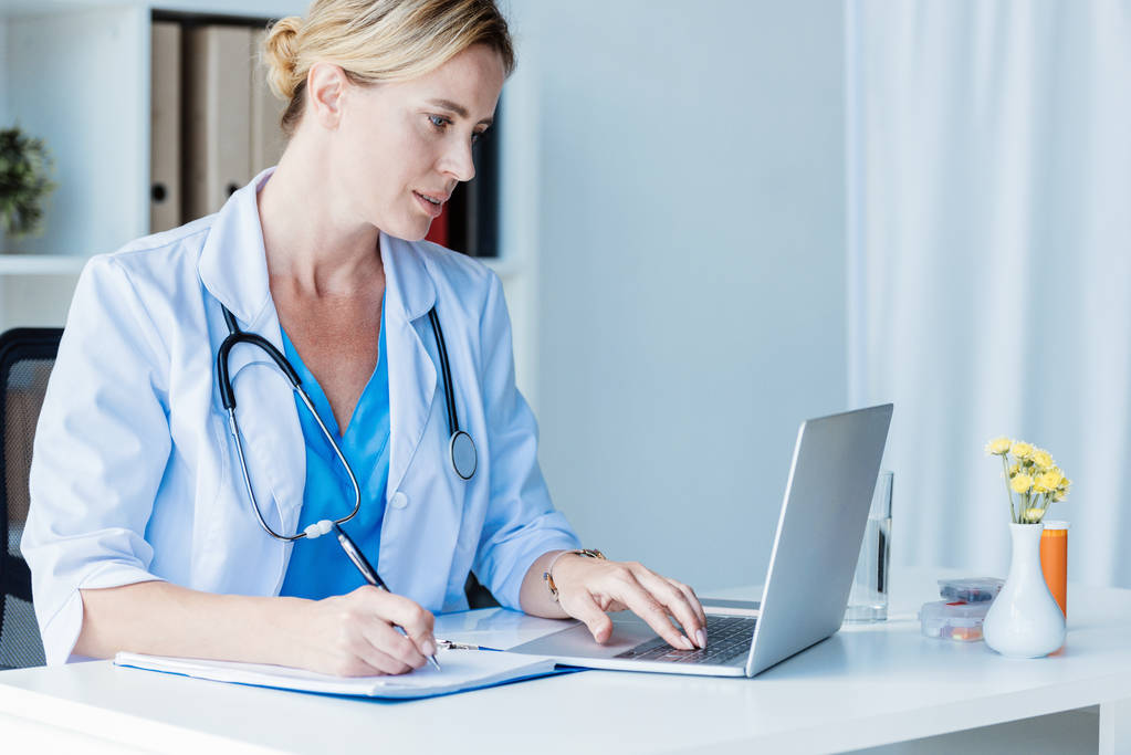 Medico donna concentrato che scrive negli appunti a tavola con laptop in ufficio
  - Foto, immagini