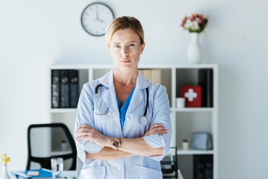 médico grave do sexo feminino em casaco branco com estetoscópio sobre o pescoço em pé com os braços cruzados olhando para a câmera no escritório
  - Foto, Imagem