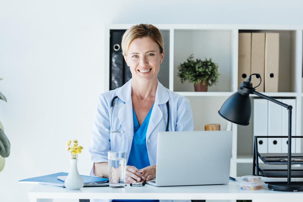 mosolygós felnőtt női orvos látszó-on fényképezőgép, és leült az asztal, laptop, iroda  - Fotó, kép