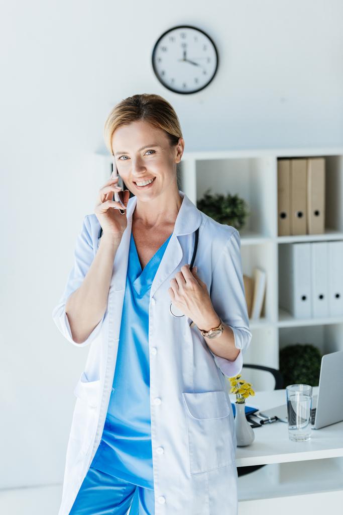 attraktive Ärztin im weißen Kittel blickt in die Kamera und spricht im Büro auf dem Smartphone  - Foto, Bild