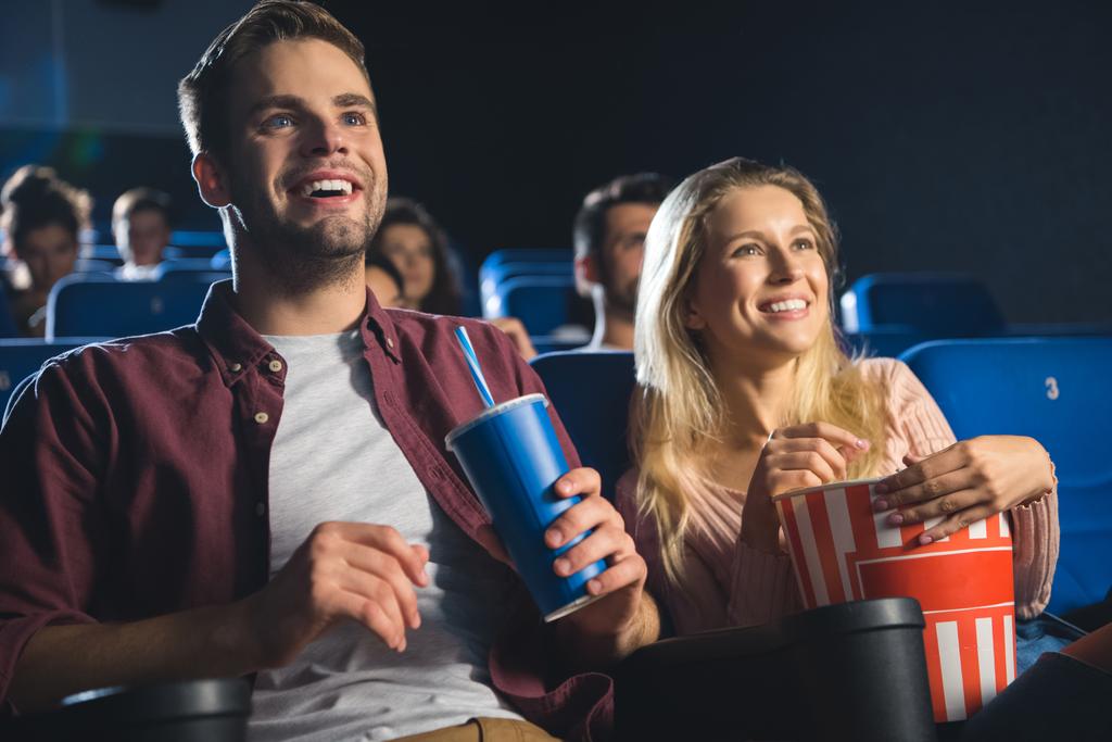 cheerful couple with popcorn and soda drink watching film together in cinema - Photo, Image