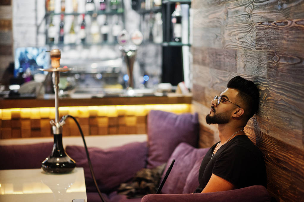Stylowe broda arabian człowiek w okularach i czarny t-shirt palenie fajki kryty bar. Emiraty modelu o odpoczynku. - Zdjęcie, obraz