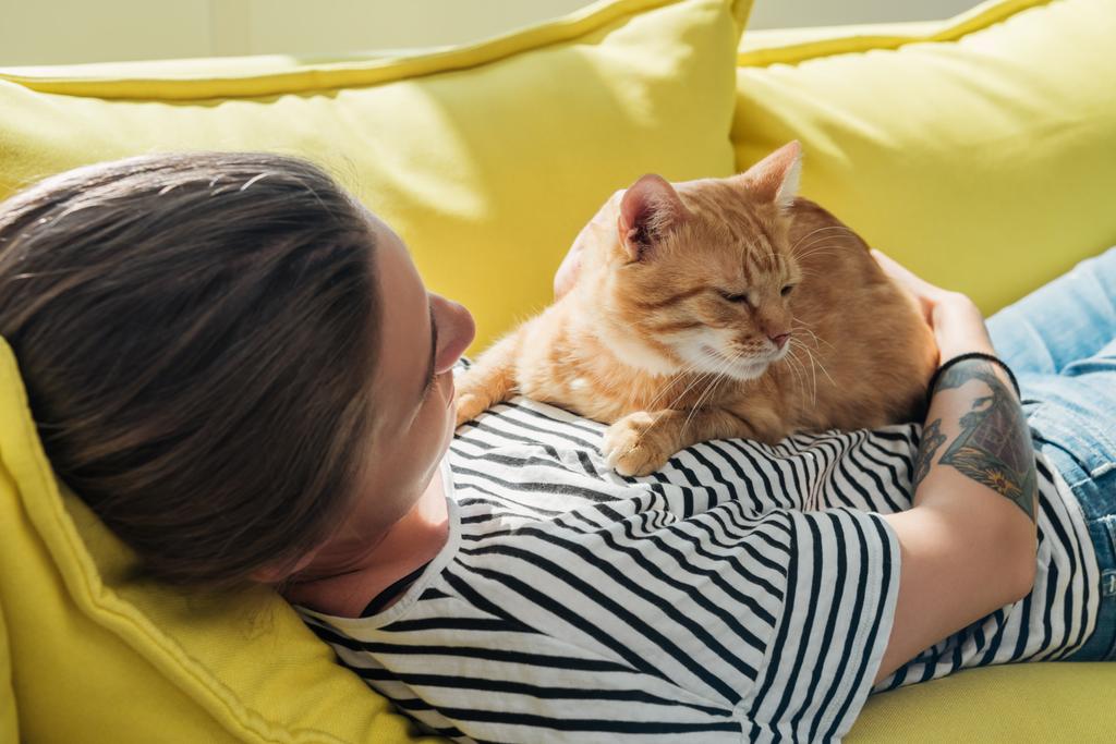 young woman holding cute red cat and lying on yellow couch - Photo, Image