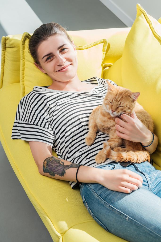 hoge hoekmening van meisje bedrijf rode kat en glimlachend in de camera terwijl liggend op gele Bank - Foto, afbeelding