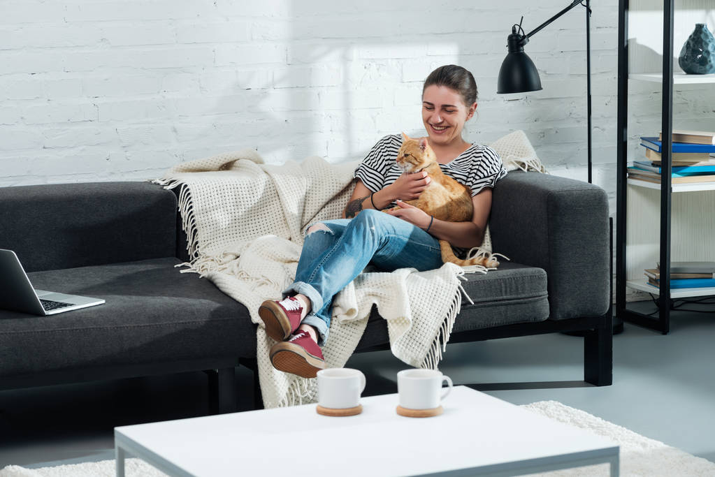 schönes lächelndes Mädchen sitzt auf dem Sofa und palmt süße Ingwerkatze im Wohnzimmer - Foto, Bild