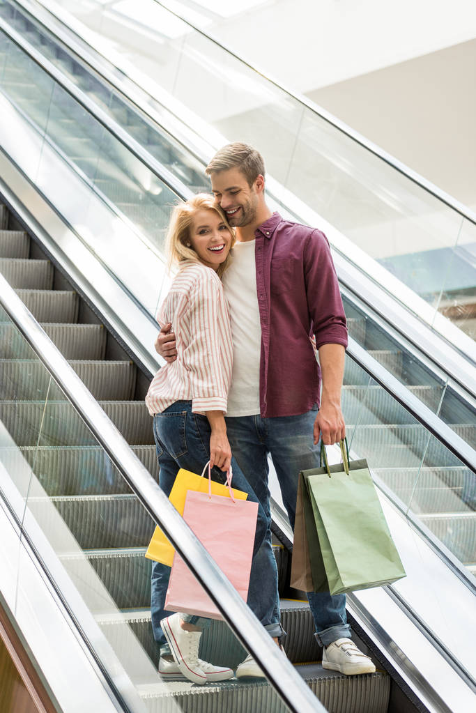 knappe Glimlachende man met boodschappentassen omarmen vriendin op roltrap bij winkelcentrum  - Foto, afbeelding