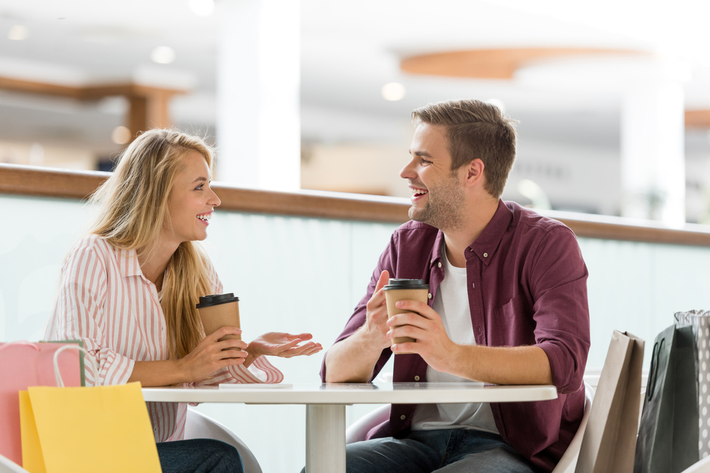 Lächelndes junges Paar mit Pappbechern Kaffee sitzt nach dem Einkauf am Tisch im Café  - Foto, Bild