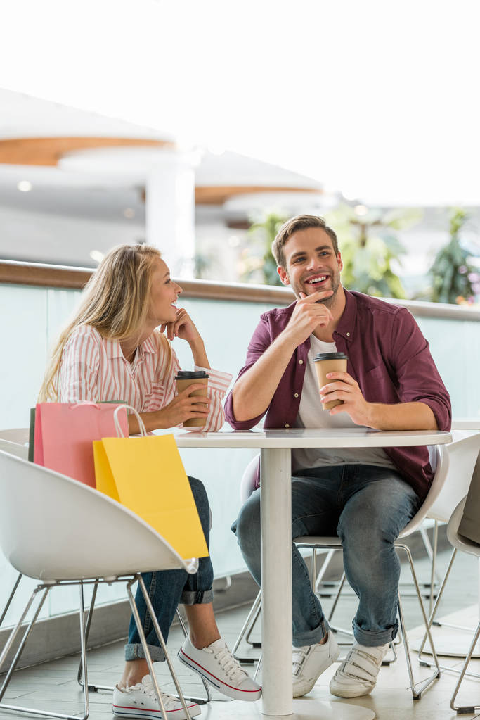  glückliches junges Paar mit Einkaufskäfern am Tisch mit Einwegbechern Kaffee im Café  - Foto, Bild