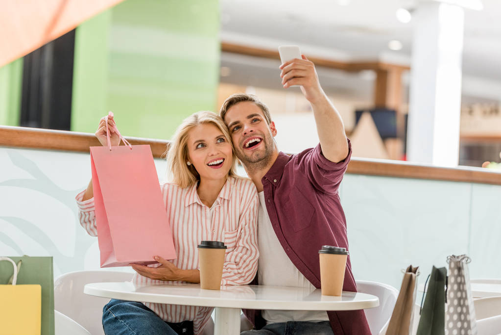 człowiek biorąc selfie z dziewczyna pokazując torba na zakupy przy stole z jednorazowych kubków kawy w kawiarni  - Zdjęcie, obraz